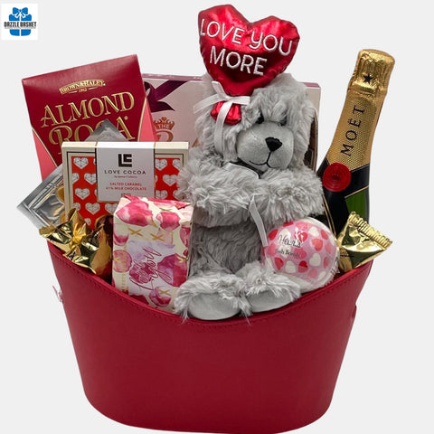A red color themed valentine gift basket that includes a bottle of champagne, "Love you More"  teddy bear and delicious gourmet snacks including cheese and chocolates made in a red faux leather oval container