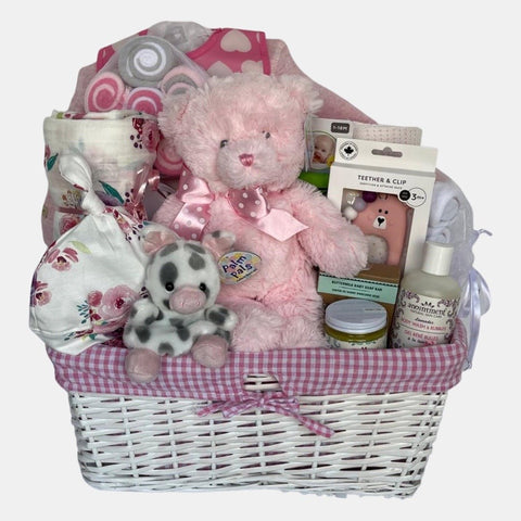 A baby girl gift basket that includes pink color themed baby products including blanket, onesie, baby care and a beautiful teddy bear packed in a lovely white basket.